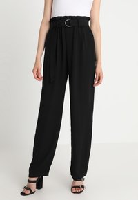 Pantalons noirs taille haute avec une ceinture dotée d'un anneau en métal, texture lisse, jambes larges et coupe décontractée. Associés à des talons à lanières.