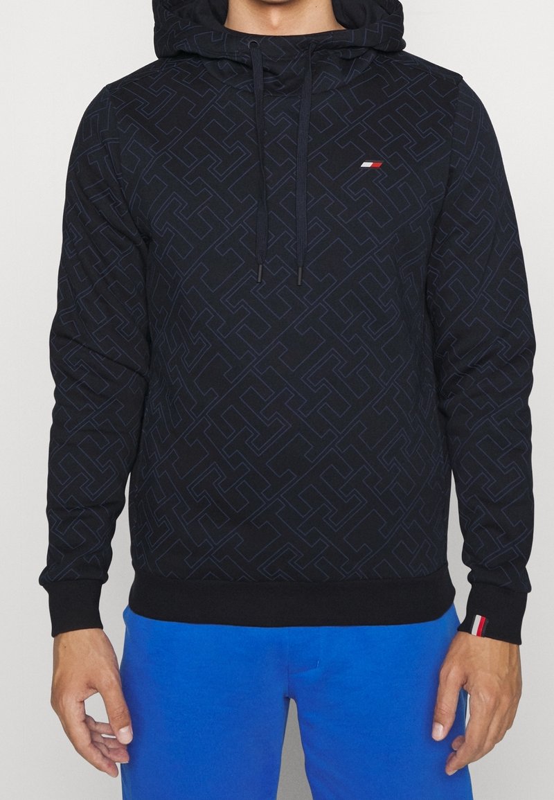 Personne portant un sweat à capuche noir à motif avec un petit logo rouge, blanc et marine et un pantalon bleu vif, les mains détendues sur les côtés.