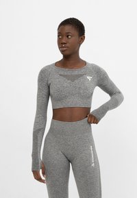 Sivý priliehavý crop top s dlhými rukávmi, ktorý má sieťkový detail na hrudníku a otvory na prsty, spárovaný s vyššími legínami z rovnakého materiálu.