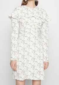 Un vestido blanco con un patrón floral que presenta flores rosas y hojas verdes. Mangas largas, cuello con volantes y detalle fruncido en la parte delantera para añadir textura.