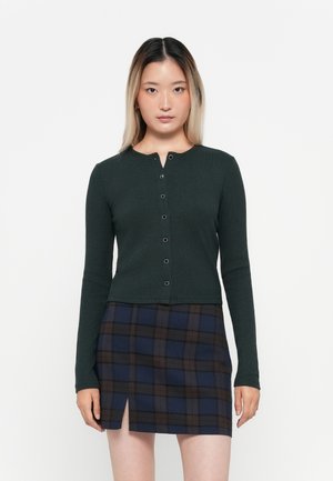 Mörkgrön ribbad långärmad cardigan med rundad halsringning och fem knappar fram. Kombinerad med en marinblå och brun rutig minikjol med en slits.