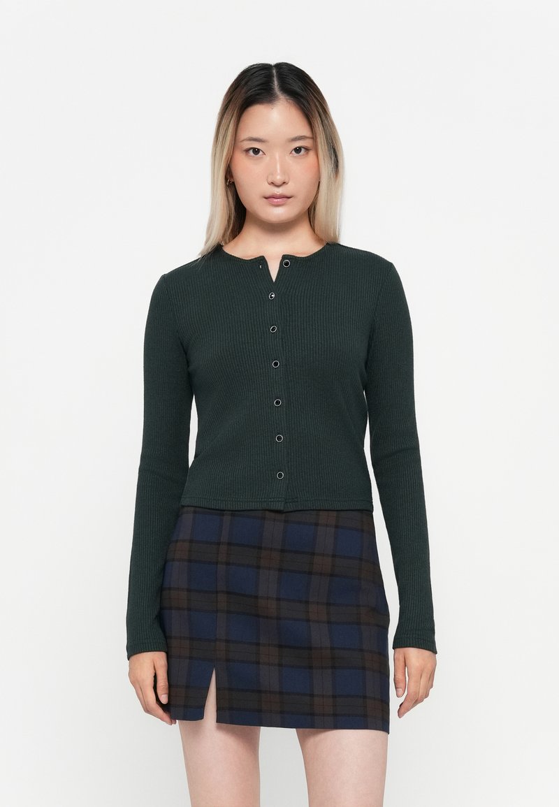 Dark green ribbed long-sleeve cardigan with a crew neckline and five front buttons. Paired with a navy and brown plaid mini skirt featuring a slit.
