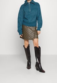 Pull en maille teal avec un col montant, mini jupe matelassée olive, et bottes en cuir noires montantes avec des bouts pointus et des talons.