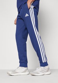 Pantalons de survêtement bleus en coton avec un design à trois bandes blanches sur les côtés, taille élastique et poignets côtelés, associés à des chaussures de sport gris clair.