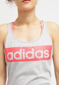 Šedý tílko s velkým červeným obdélníkovým grafickým prvkem, na kterém je bílé písmo "adidas". Design zahrnuje zakřivený výstřih a tenké ramínka.