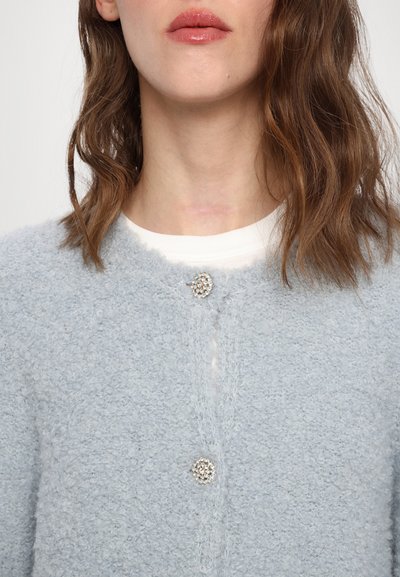 Lys blå cardigan laget av teksturert stoff, med rund halslinning og en dekorativ knapp med krystalldesign i midten.