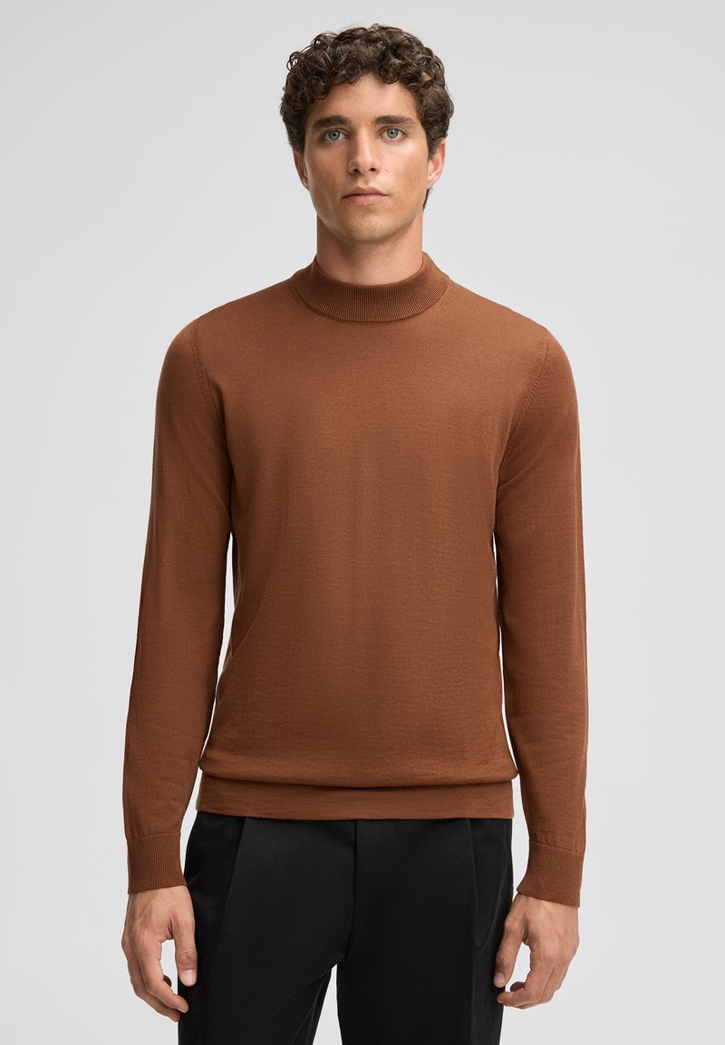 Ein Mann steht vor einem neutralen Hintergrund, trägt einen braunen Rollkragenpullover und schwarze Hosen und hat einen ernsten Gesichtsausdruck.