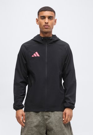Mężczyzna w czarnej kurtce z kapturem na zamek błyskawiczny z różowym logo Adidas na lewej piersi i szarych spodniach, stojący na jednolitym tle.
