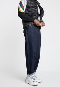 Gilet imbottito nero sopra una maglietta a maniche lunghe grigia con strisce colorate, abbinato a pantaloni larghi in denim scuro e sneakers bianche.