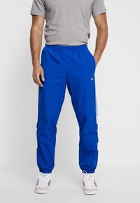 Blå träningsbyxor i lätt tyg, med elastisk midja, sidofickor och vita ränder längs benen.