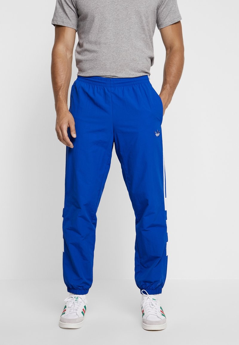 Blå träningsbyxor i lätt tyg, med elastisk midja, sidofickor och vita ränder längs benen.