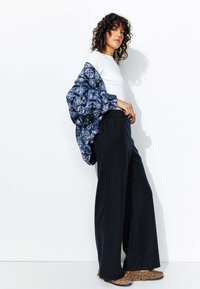 Veste matelassée à motif bleu sur un haut blanc, associée à un pantalon large noir et des chaussures à imprimé léopard. Textures lisses et couleurs vives.