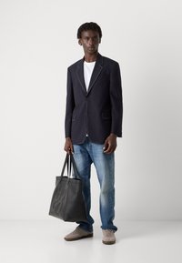 Blazer azul-marinho com bainhas brancas, combinado com uma t-shirt branca e jeans azul-claro. Segurando uma bolsa tote de couro preta, usando ténis cinzentos.