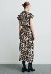 Bruine maxi-jurk met bloemmotief, korte mouwen en een taille met riem. Gecombineerd met zwarte, kniehoge enkellaarsjes met hak, afgebeeld tegen een effen achtergrond.