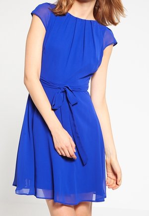 Mujer con un vestido azul real hasta la rodilla, de mangas cortas transparentes y cinturón atado a la cintura, posando frente a un fondo claro.