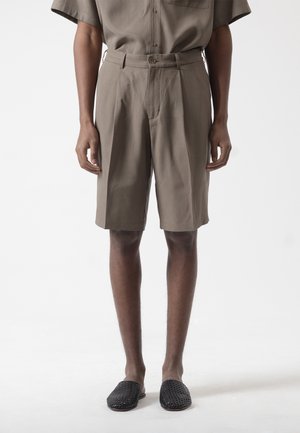 Hombre con pantalones cortos marrones de tiro alto hasta la rodilla con pliegues, combinados con camisa de manga corta a juego y zapatos negros tejidos sin cordones.