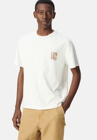 Camiseta de algodón blanca con un pequeño estampado marrón "LDN PRS" en el lado izquierdo del pecho. Mangas cortas y un cuello redondo clásico.