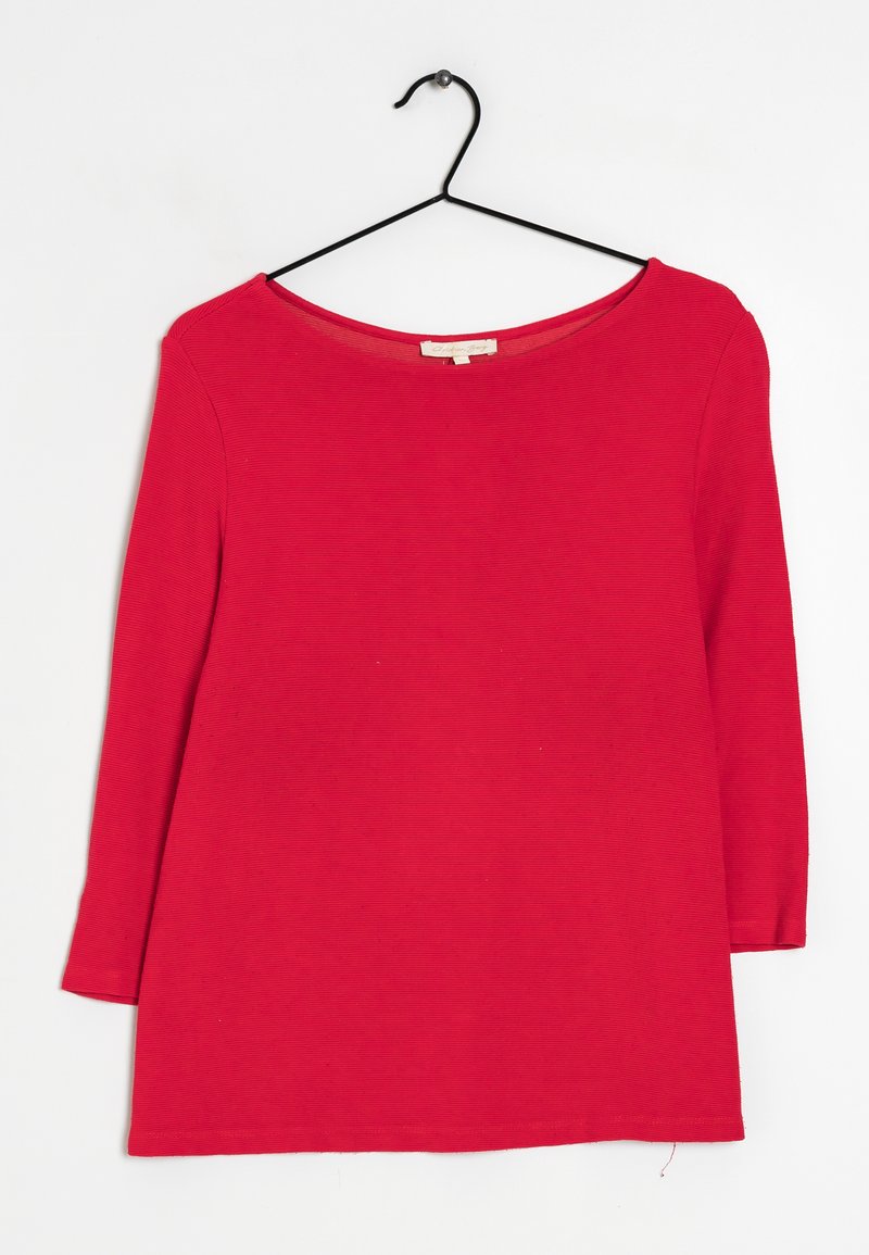 Top rouge à manches longues avec un col rond, en tissu côtelé. Présente une coupe droite et une étiquette au dos du col. Accroché à un crochet noir.
