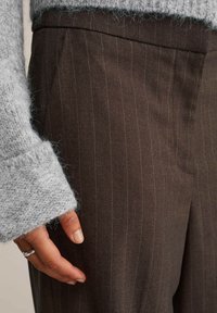Ezüst gyűrűt viselő kéz barna csíkos nadrág mellett, szürke kötött pulcsi ujjával részben eltakarva a csuklót.