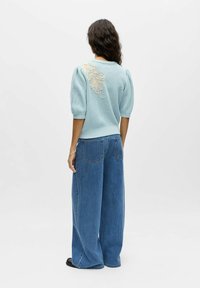 Pull bleu tricoté avec manches bouffantes et une broderie beige décorative dans le dos, associé à un jean en denim à jambes larges.