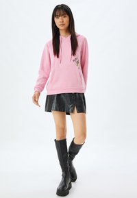 Sudadera con capucha rosa, con un bolsillo frontal y un detalle bordado que dice "Hola", combinada con una minifalda de cuero sintético negra y botas hasta la rodilla.