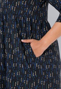 Hand rustend in zijvak van zwarte jurk met blauw, beige en teal verticale stippenpatroon.