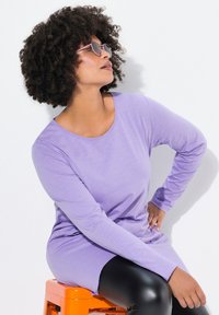 Lila Langarm-Oberteil aus weichem Stoff, mit einem runden Halsausschnitt und lässiger Passform. Getragen mit glänzenden schwarzen Leggings.