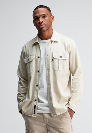 Beige overhemd met lange mouwen, twee borstzakken, zwarte knopen en een casual pasvorm, gecombineerd met een witte T-shirt eronder en beige broek.