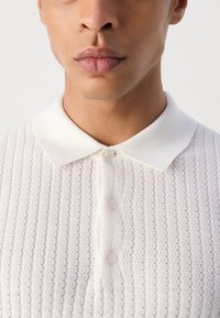 Witte gebreide polo met een gestructureerd patroon, een kraag en drie knopen aan de voorkant voor sluiting.