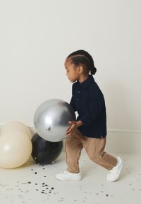 Polo bleu marine, pantalon beige et baskets blanches. Enfant tenant un ballon argenté brillant. Ballons en arrière-plan : noir, beige et argenté.