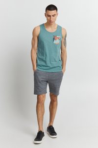 Tyrkysový tielko s vzorovaným vreckom, doplnený o sivé šortky. Oblečenie má voľný strih a základné čierne tenisky.