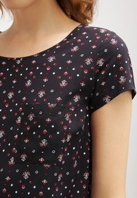 Černý top s krátkým rukávem a kulatým výstřihem, který má květinový potisk v červené, bílé a zelené barvě. Obsahuje malou náprsní kapsu. Hladká textura látky.