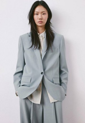 Young woman with long black hair wearing oversized light gray suit jacket and pants, striped button-up shirt, hands in pockets, neutral expression.