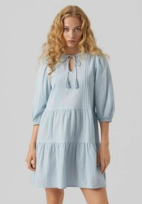 Robe bleu clair avec une jupe à volants, un col en forme de clé, des détails à nouer et des manches bouffantes. Fabriquée en tissu texturé.