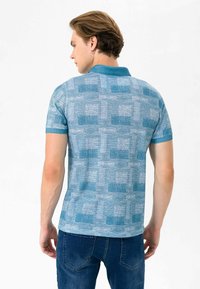 Kortærmet polo-shirt i lyseblå med et struktureret mønster, der har en ensfarvet krave og subtile rektangulære detaljer. Bæres med mørk denim jeans.