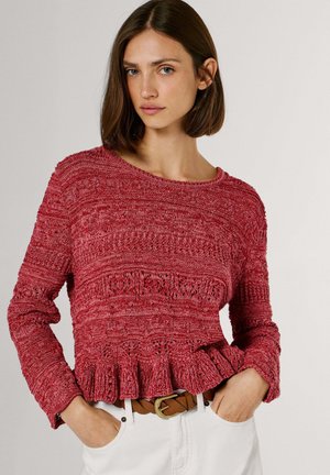 Frau mit kurzem braunem Haar, die einen strukturierten roten Strickpullover mit gerafftem Saum und weiße Hosen mit einem braunen geflochtenen Gürtel trägt.