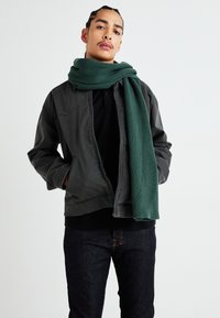 Casaco cinza com fecho na frente e bolsos laterais, combinado com um cachecol verde às riscas. Camisa preta e jeans escuros completam o visual.