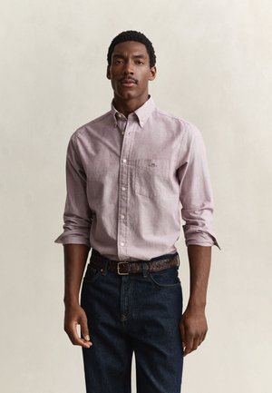Mann trägt ein helllila Hemd mit Knöpfen und hochgekrempelten Ärmeln, dunkle Jeans und einen geflochtenen braunen Gürtel und steht vor einem schlichten beigen Hintergrund.