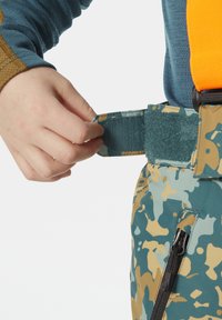 Kolorowe spodnie w kamuflaż z grubym pomarańczowym paskiem i zielonym rzepem. Ręka dostosowuje rzep przy pasie.