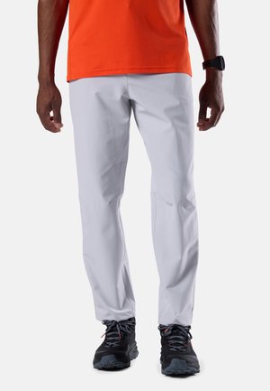 Mann trägt hellgraue Hose, schwarze Wanderschuhe mit grauen Schnürsenkeln, orangefarbenes Hemd und schwarze Armbanduhr, steht vor weißem Hintergrund.