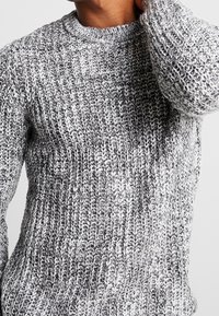 Grau und weißer gestrickter Pullover mit Rundhalsausschnitt, der ein strukturiertes Design und sichtbare Nähdetails aufweist.