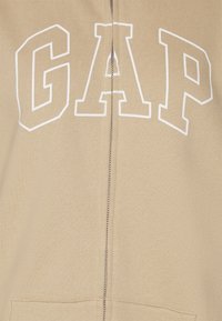 Sudadera con capucha beige con cremallera y "GAP" en letras blancas, con tejido texturizado y bolsillos delanteros. La cremallera corre verticalmente por el centro.
