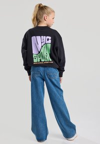 Le sweat à capuche noir présente un graphique audacieux "NYC SPORT" multicolore dans le dos. Associé à un jean en denim bleu clair à jambe large.