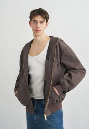 Νέος άνδρας που φορά καφέ φούτερ με φερμουάρ πάνω από λευκή φανέλα και μπλε τζιν, στέκεται με τα χέρια στις τσέπες μπροστά σε απλό φόντο.