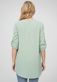 Top verde chiaro a maniche lunghe con polsini arrotolati e orlo arrotondato. Tessuto testurizzato, vestibilità casual, superficie liscia senza ulteriori motivi.