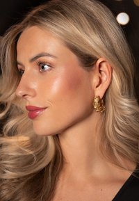 Guldøreringe med en tekstureret blomsterdesign, der har en glat, reflekterende overflade og en mellemstor størrelse, der omfavner øreflippen.