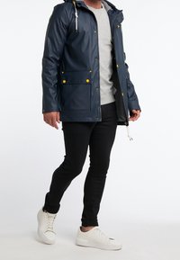 Mörkblå vattentät jacka med huva, gula knappar, två framfickor och en grå sweatshirt under, tillsammans med svarta jeans och vita sneakers.