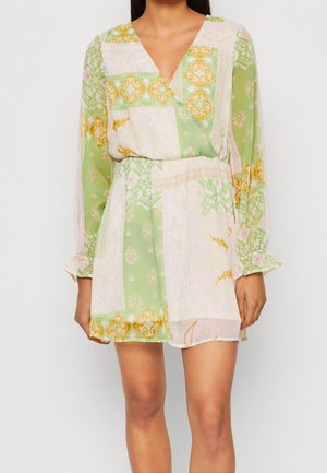 Femme portant une mini-robe à manches longues et col en V, avec des motifs floraux et géométriques verts, roses et jaunes.