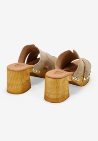 Zuecos de gamuza beige con un tacón de bloque de madera, que presentan dos correas anchas y remaches dorados en el lateral, con una plantilla acolchada beige.