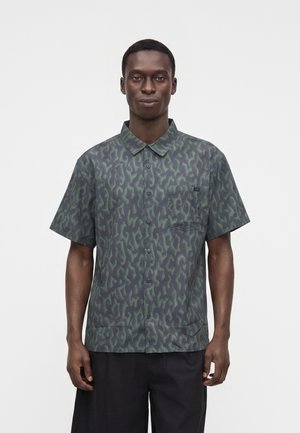Hombre con camisa de manga corta con un patrón abstracto verde y oscuro y pantalones negros, de pie frente a un fondo liso.